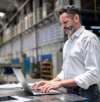 Man in a white shirt working on a laptop in an industrial factory setting.