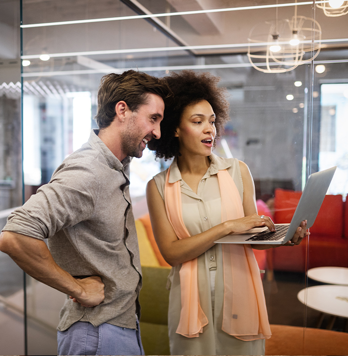 Two colleagues standing in an office, smiling and looking at a laptop together