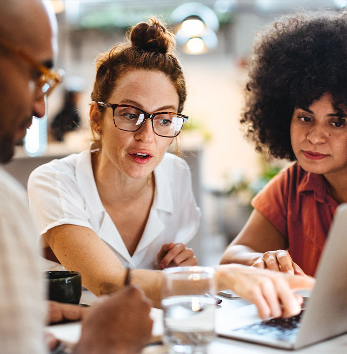 Three colleagues collaborating around a laptop, with one woman pointing at the screen and others attentively listening